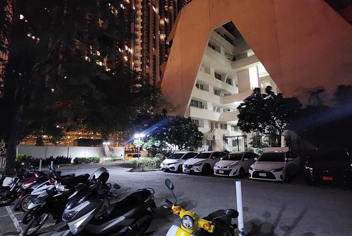 Night scene of a parking lot filled with motorcycles; a bright yellow scooter is in the foreground with a beige angular building in the background urban lighting at night.