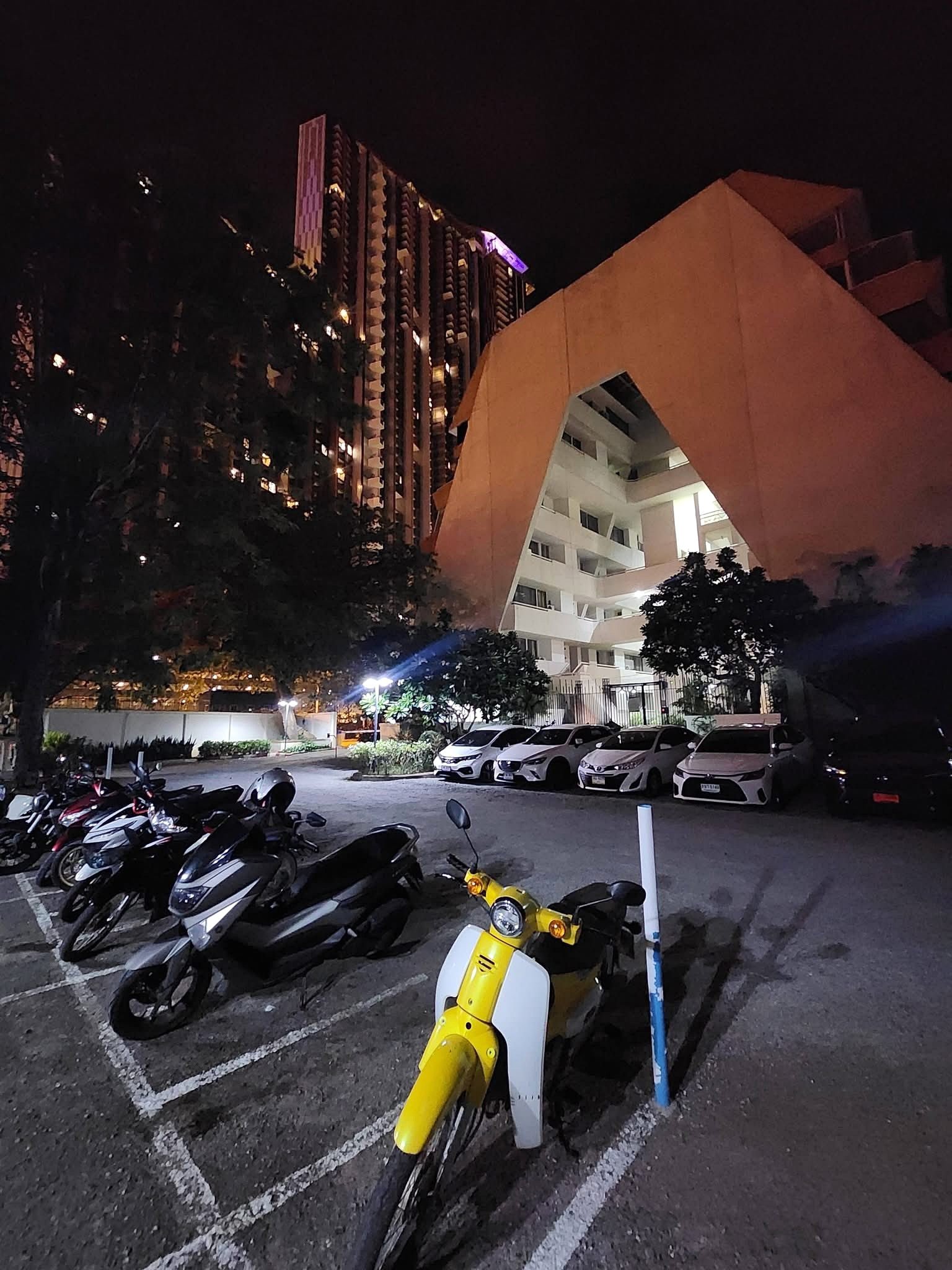 PANCHALEA_RESIDENCE_Jomtien_Rdl290_25 Night scene of a parking lot filled with motorcycles; a bright yellow scooter is in the foreground with a beige angular building in the background urban lighting at night.
