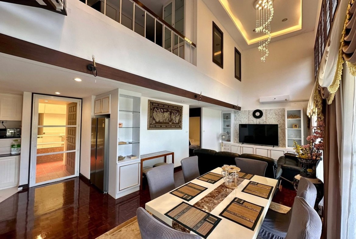 Open-concept living space with a long dining table, gray upholstered chairs, and a hanging crystal chandelier above; a second-floor balcony is visible. (Interior home scene)