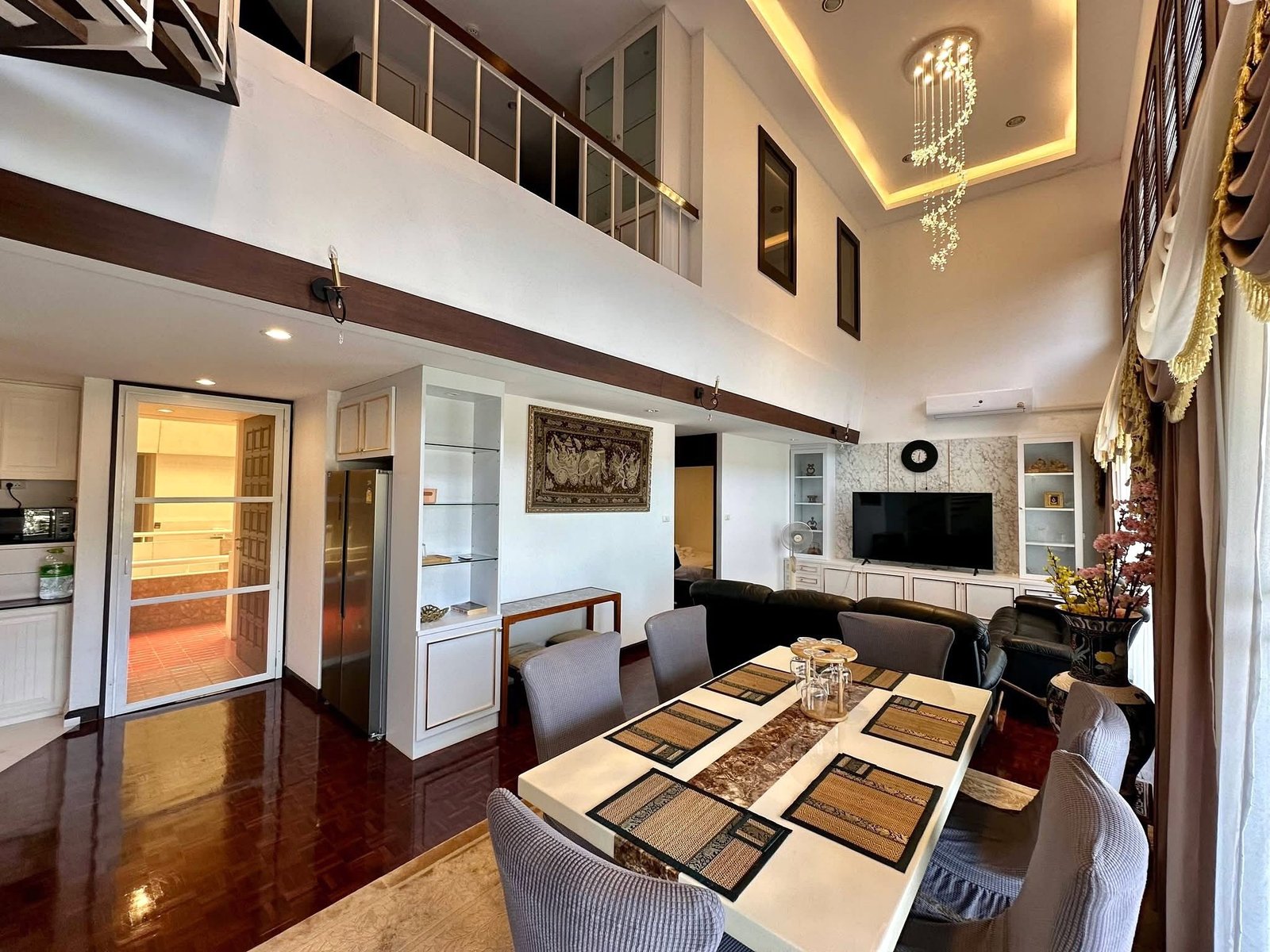 PANCHALEA_RESIDENCE_Jomtien_Rdl290_3 Open-concept living space with a long dining table, gray upholstered chairs, and a hanging crystal chandelier above; a second-floor balcony is visible. (Interior home scene)