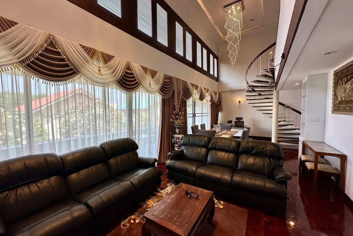 Living room with dark leather sofas, a wooden coffee table, tall windows with ornate draped curtains, and a crystal chandelier overhead.