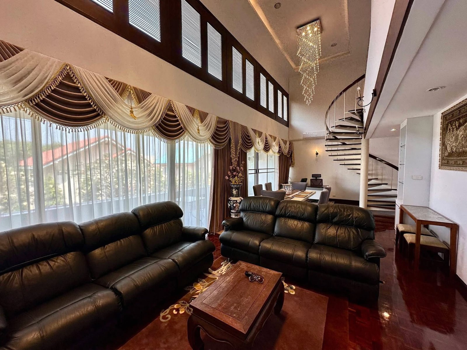 PANCHALEA_RESIDENCE_Jomtien_Rdl290_4 Living room with dark leather sofas, a wooden coffee table, tall windows with ornate draped curtains, and a crystal chandelier overhead.