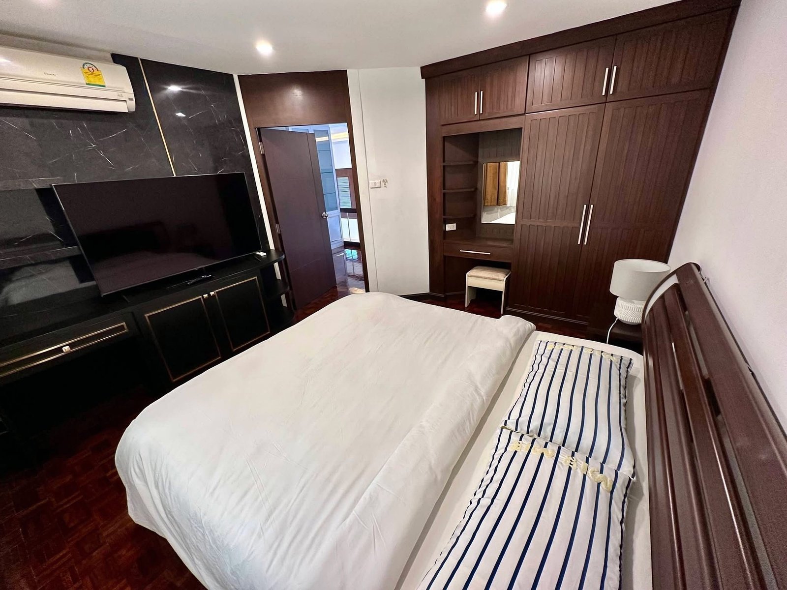 PANCHALEA_RESIDENCE_Jomtien_Rdl290_6 Twin beds with white duvets and navy striped pillows in a wood-furnished hotel-style bedroom with a flat-screen TV and dark wardrobe.