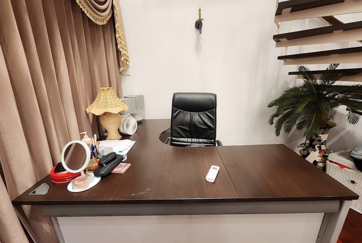 Office desk with a black leather chair, desk items, and a plant in a corner, against beige curtains and a white wall.
