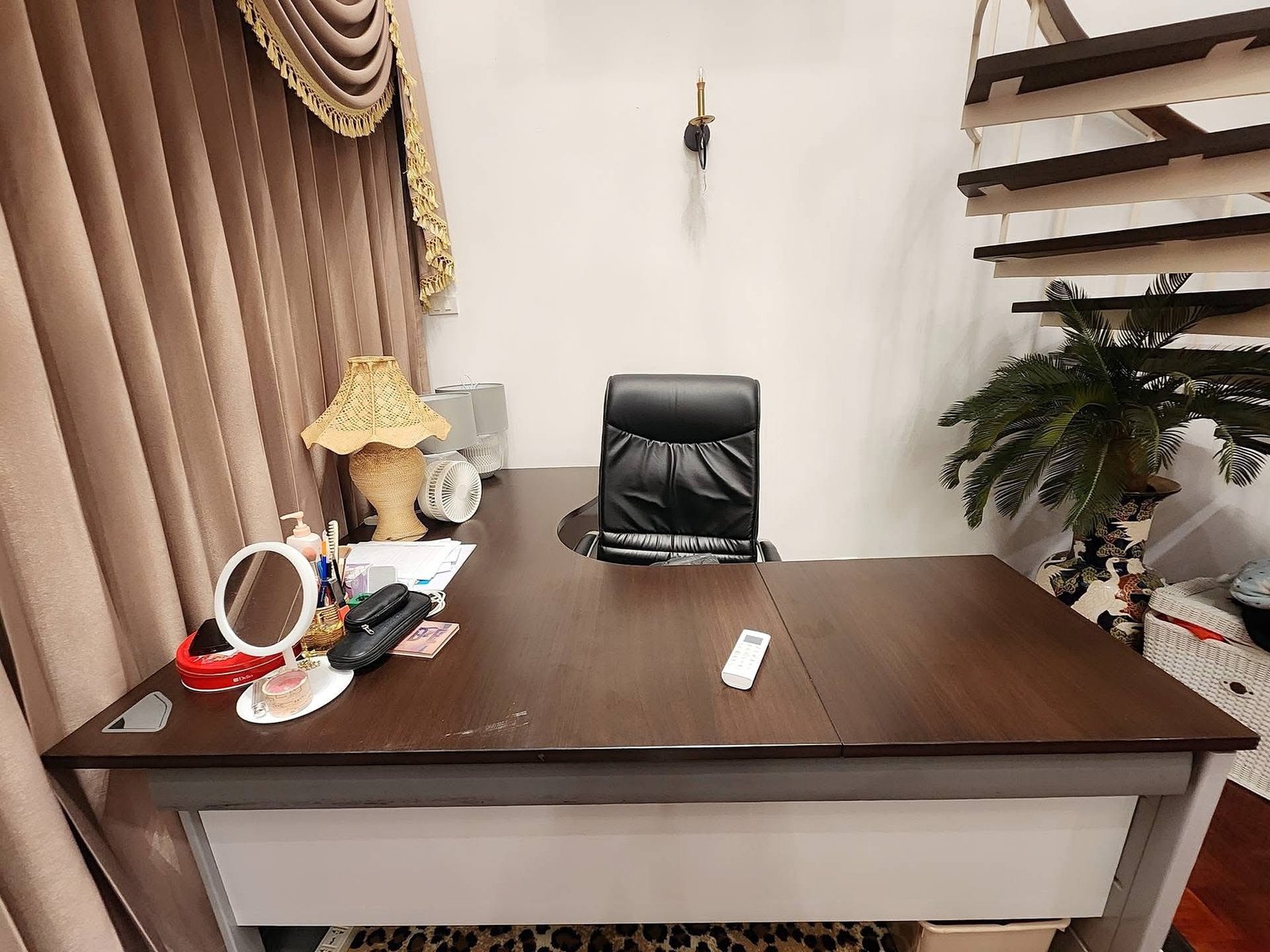 PANCHALEA_RESIDENCE_Jomtien_Rdl290_7 Office desk with a black leather chair, desk items, and a plant in a corner, against beige curtains and a white wall.