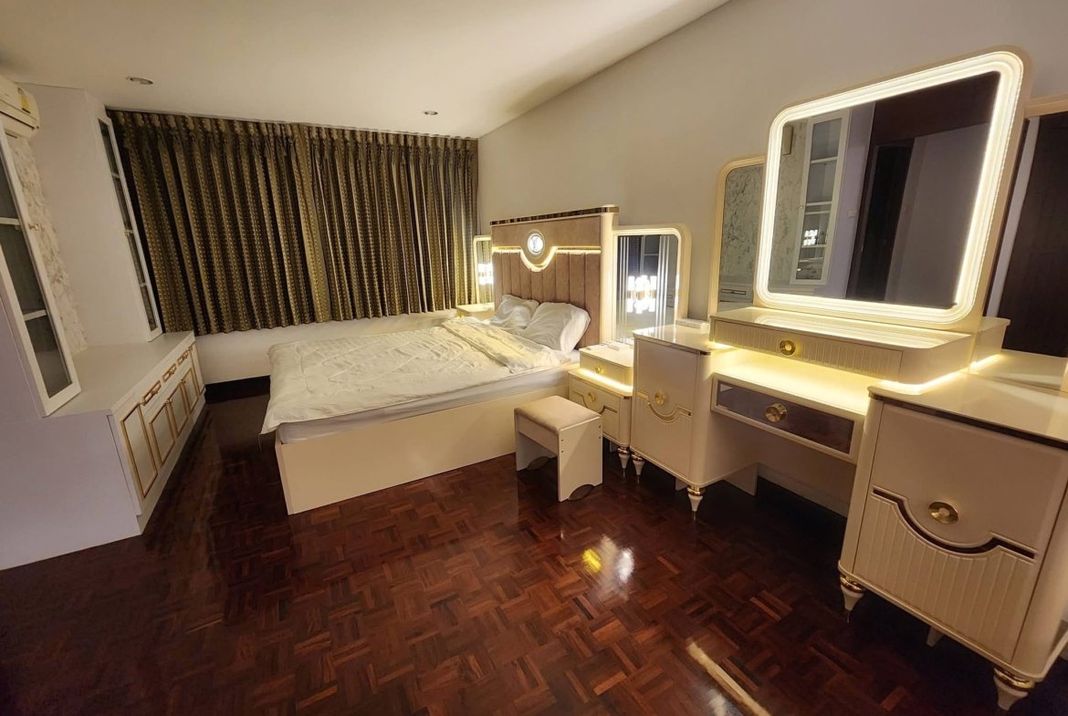 Hotel bedroom with a tufted headboard, white vanity and illuminated mirror, and long patterned drapes along a large window.