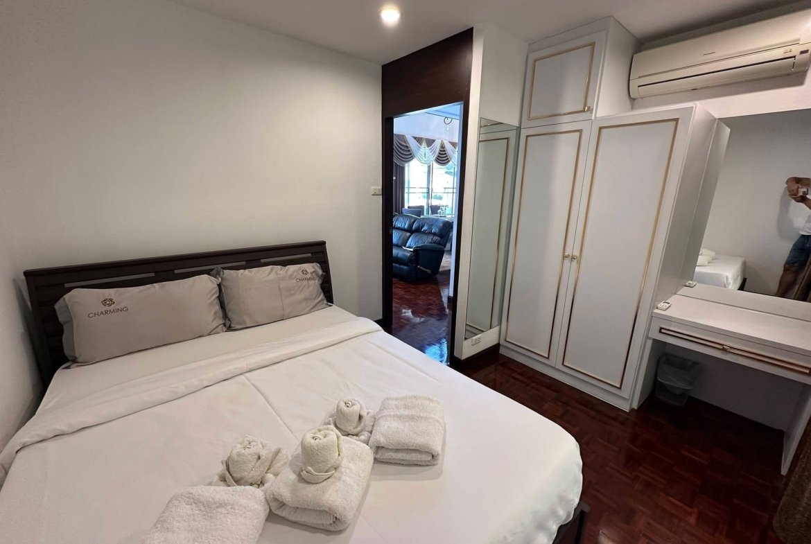 Hotel-style bedroom with a white bed, two gray pillows marked 'CHARMING', and rolled towels at the foot of the bed, plus a dark wood headboard and mirrored wardrobe doors.