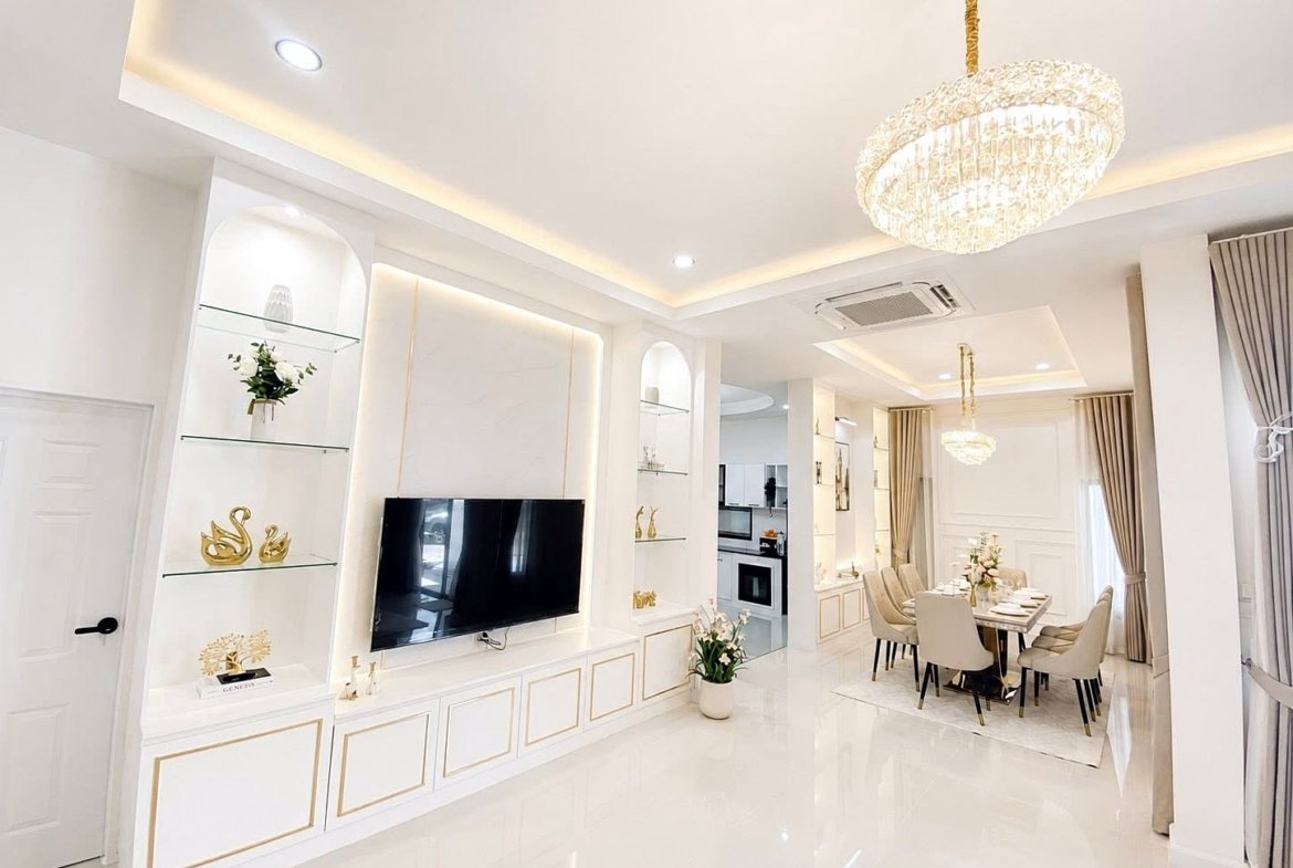 Bright open-concept living and dining area with a wall-mounted TV, white built-in cabinets, glass shelves, and a crystal chandelier.