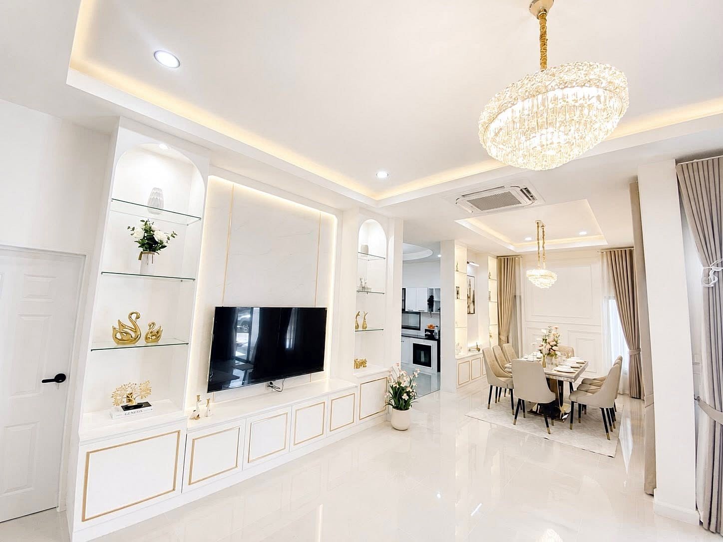 Bright open-concept living and dining area with a wall-mounted TV, white built-in cabinets, glass shelves, and a crystal chandelier.