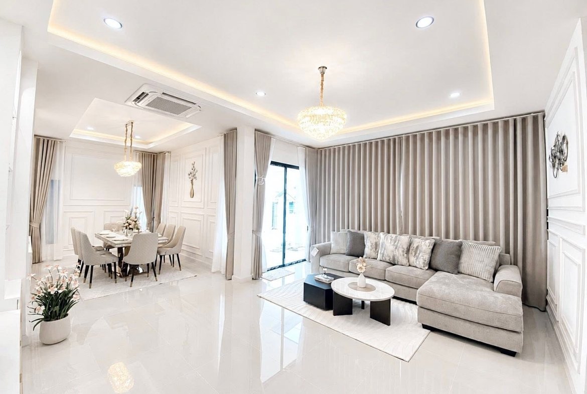 Bright open-concept living and dining area with a gray sectional sofa, marble coffee table, and floor-to-ceiling beige curtains under crystal chandeliers.