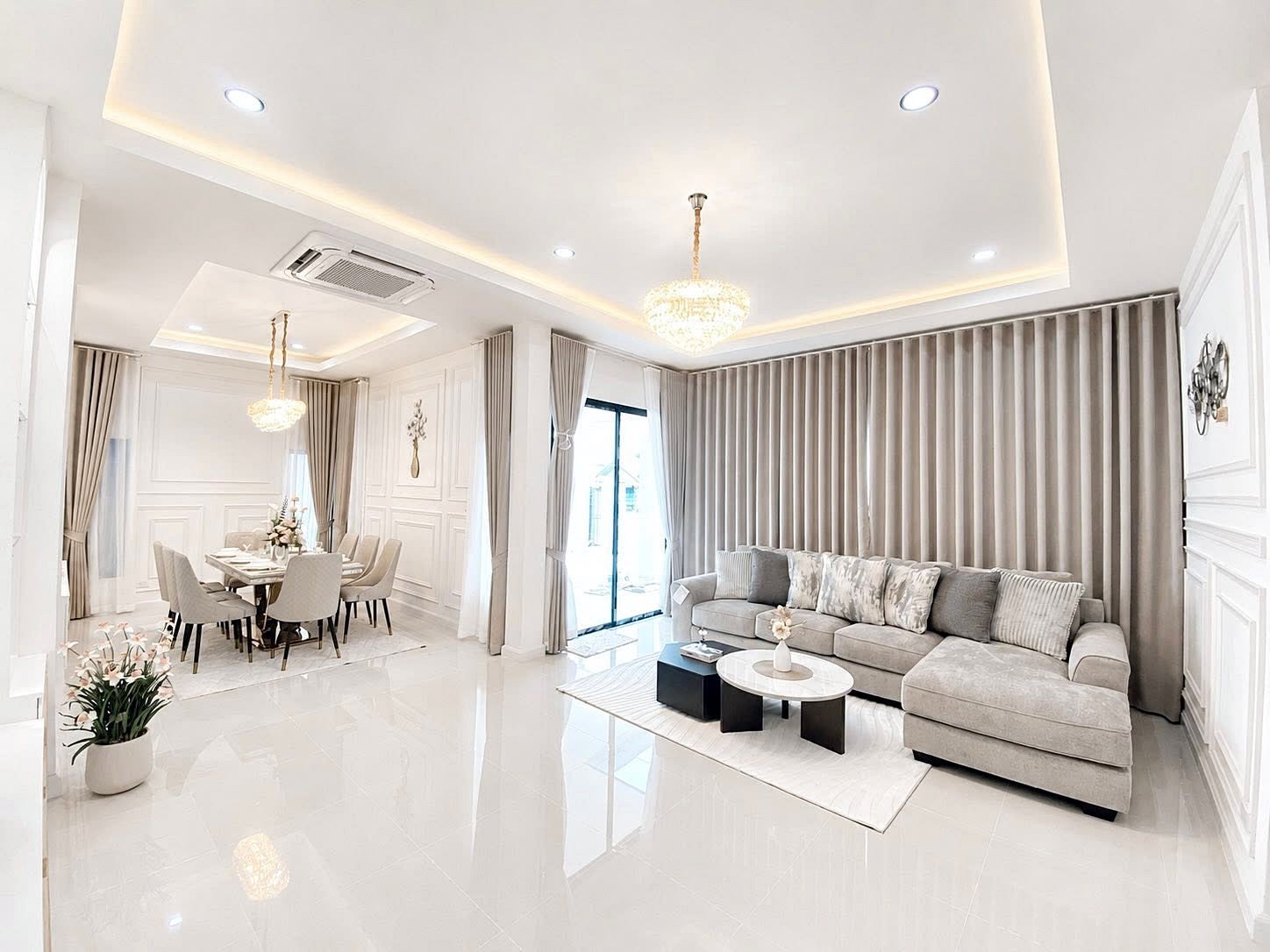 Bright open-concept living and dining area with a gray sectional sofa, marble coffee table, and floor-to-ceiling beige curtains under crystal chandeliers.