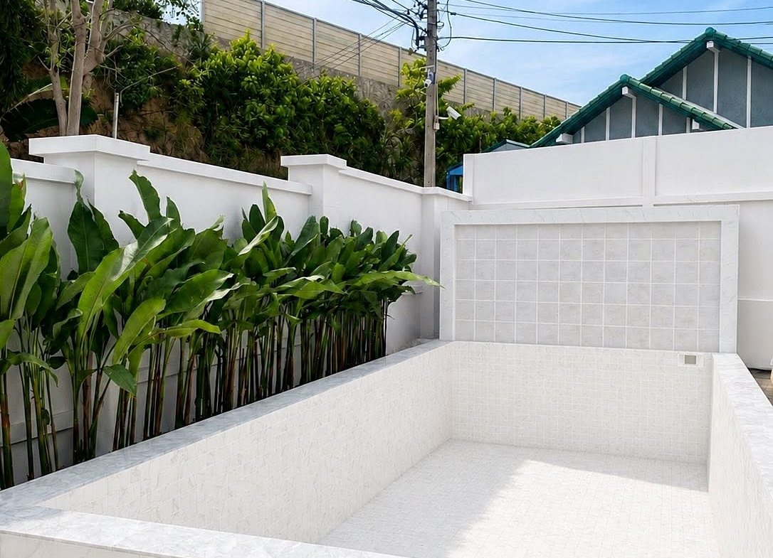 Empty white-tiled rectangular pool with a surrounding white wall and tall green plants on the left.