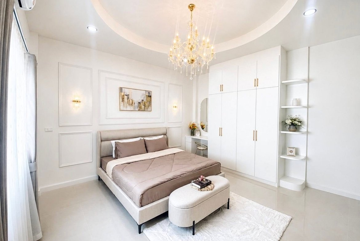 Bright white bedroom with beige upholstered bed, matching bench, and a large built-in wardrobe under a gold chandelier.