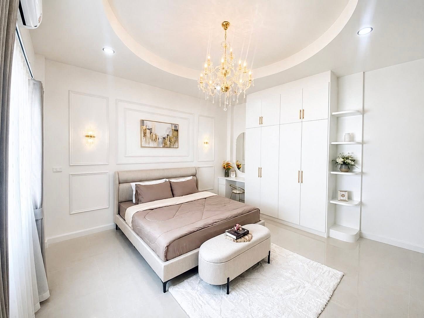 Bright white bedroom with beige upholstered bed, matching bench, and a large built-in wardrobe under a gold chandelier.