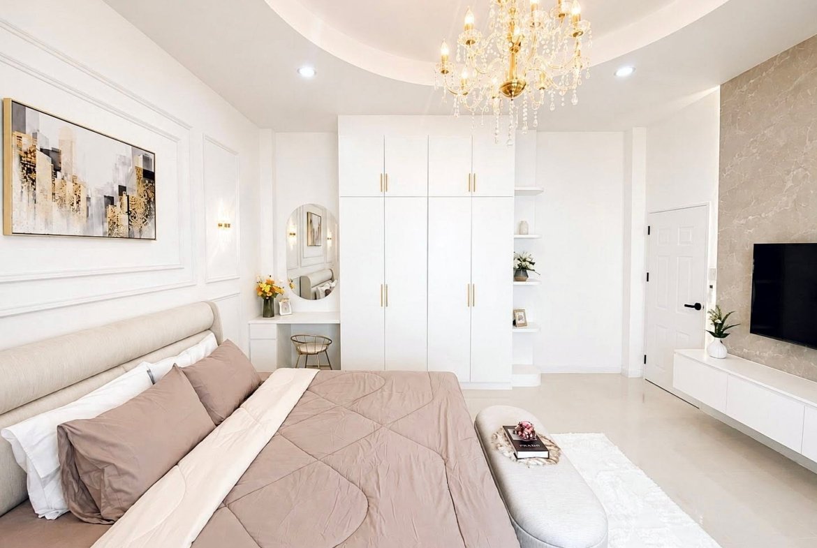 Bright bedroom with white and beige decor, chandelier, large bed, and a wall-mounted TV.