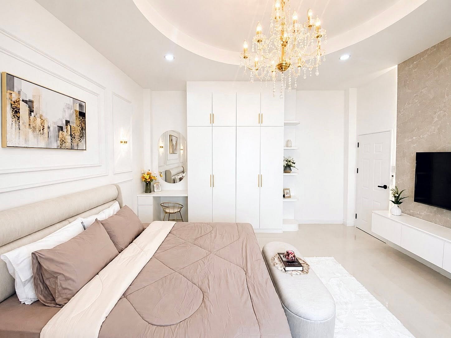 Bright bedroom with white and beige decor, chandelier, large bed, and a wall-mounted TV.