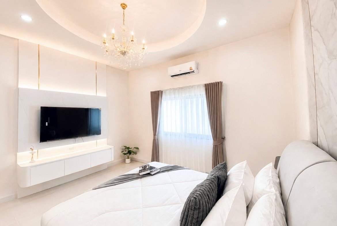 Bright modern bedroom with a crystal chandelier, a wall-mounted TV, and a white media console beneath; bed with white linens and gray pillows in the foreground.