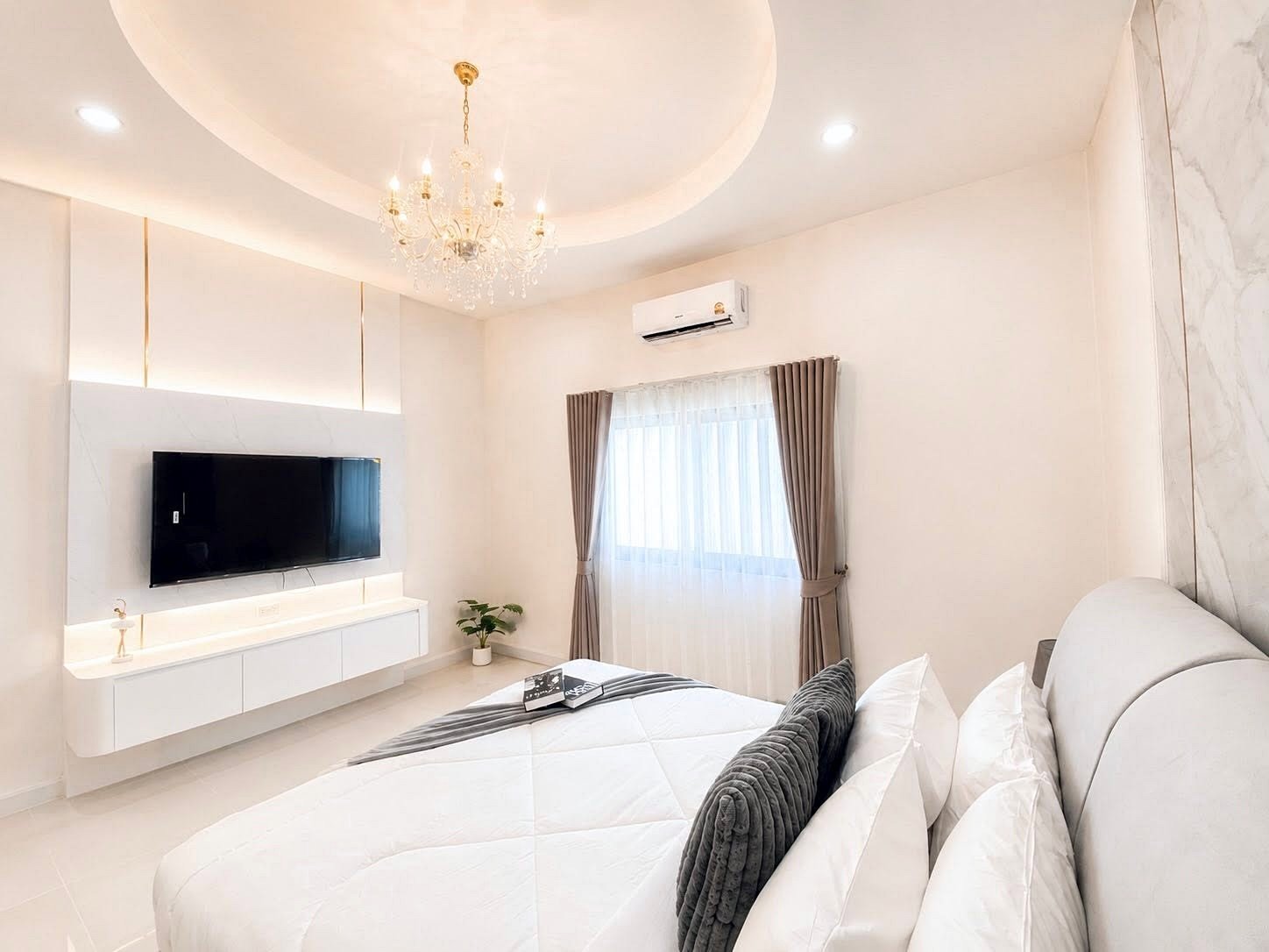 Bright modern bedroom with a crystal chandelier, a wall-mounted TV, and a white media console beneath; bed with white linens and gray pillows in the foreground.