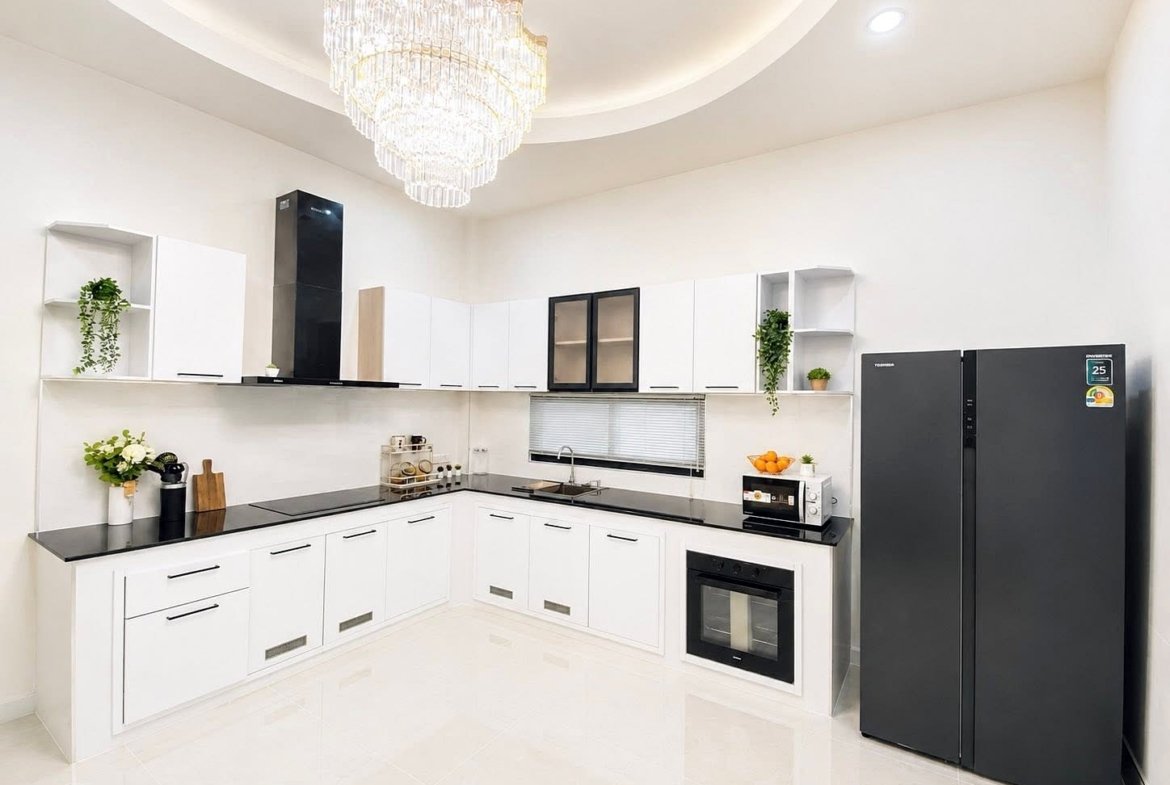 Bright modern L-shaped kitchen with white cabinets and black countertops, a large crystal chandelier, stainless steel oven and microwave, and a dark refrigerator; plants on shelves add a touch of greenery.