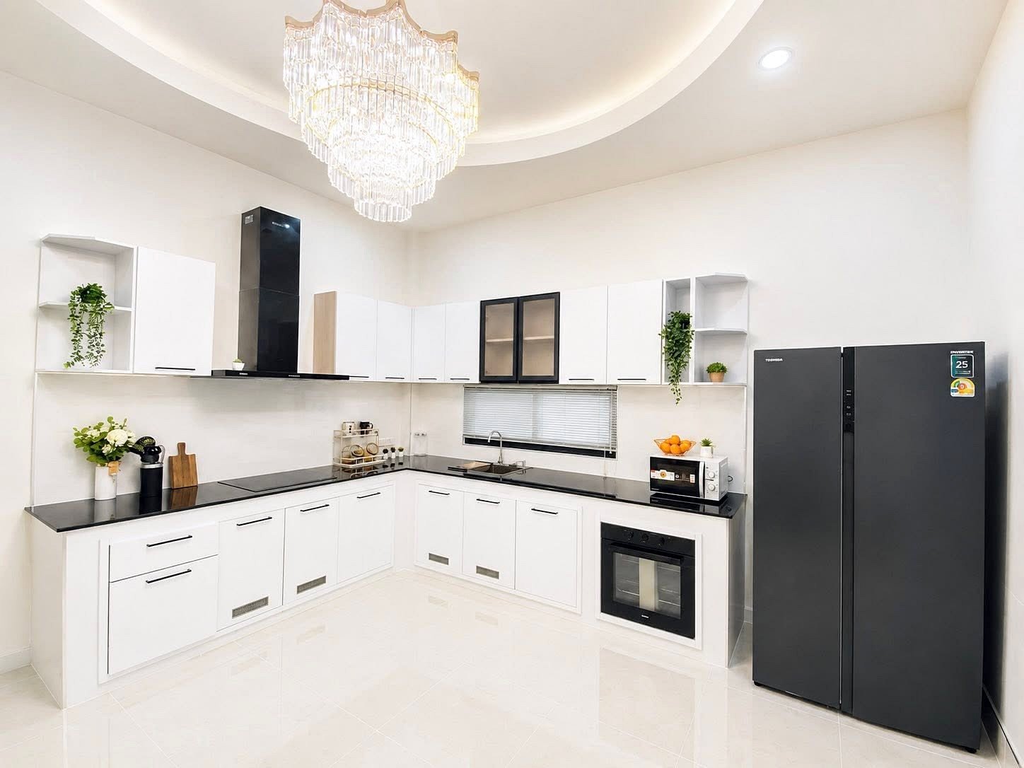 Bright modern L-shaped kitchen with white cabinets and black countertops, a large crystal chandelier, stainless steel oven and microwave, and a dark refrigerator; plants on shelves add a touch of greenery.
