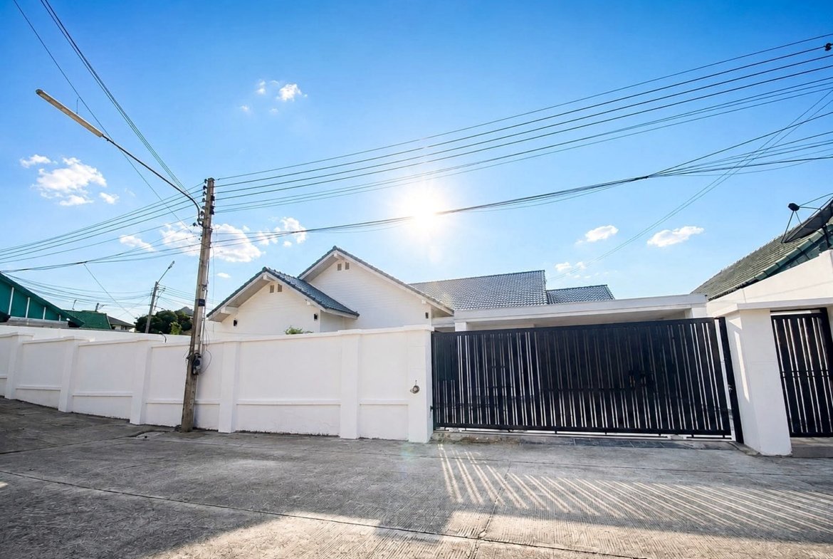 Sunny residential street with white walls, a tall black gate, and overhead power lines against a blue sky with bright sun.