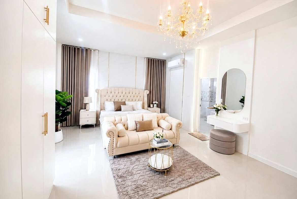 Luxurious white bedroom with a tufted beige bed, matching sofa, chandelier, and polished floors each reflecting light.