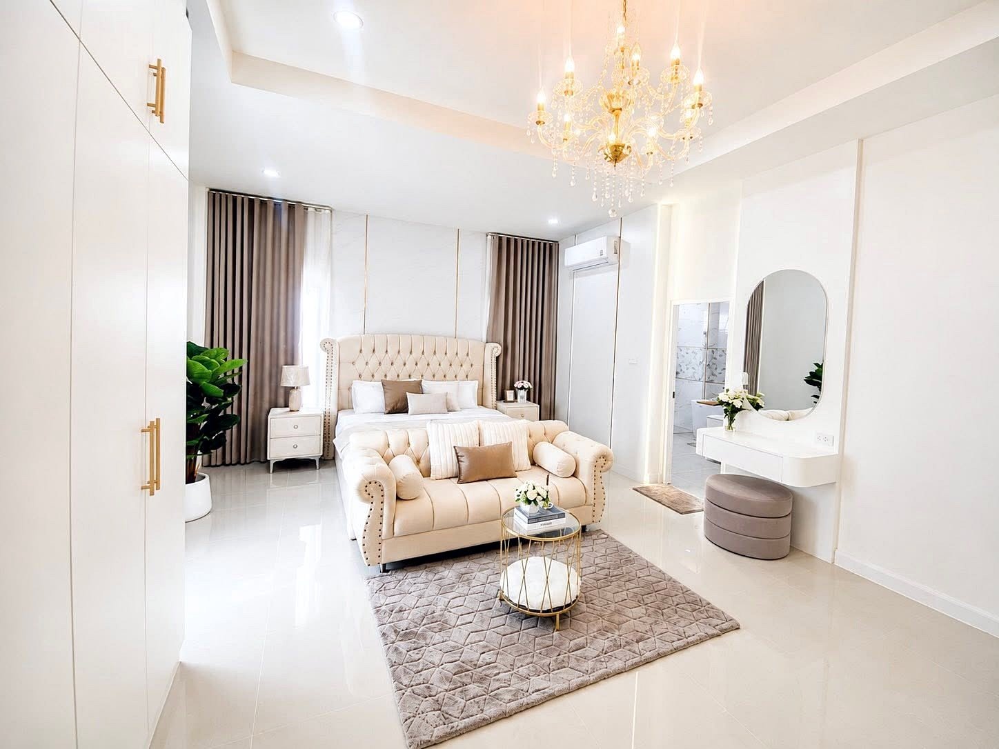 Luxurious white bedroom with a tufted beige bed, matching sofa, chandelier, and polished floors each reflecting light.