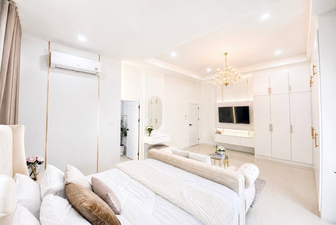 Bright white luxury bedroom with a tufted bed, chandelier, and a wall-mounted TV in an open-plan space.