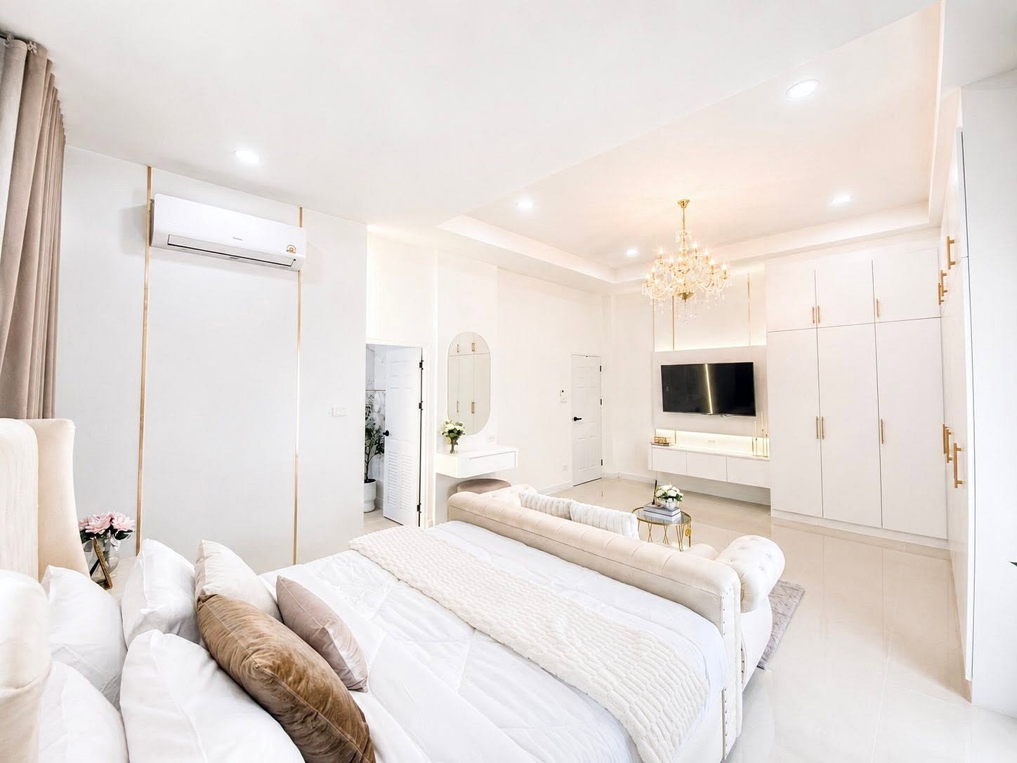 Bright white luxury bedroom with a tufted bed, chandelier, and a wall-mounted TV in an open-plan space.