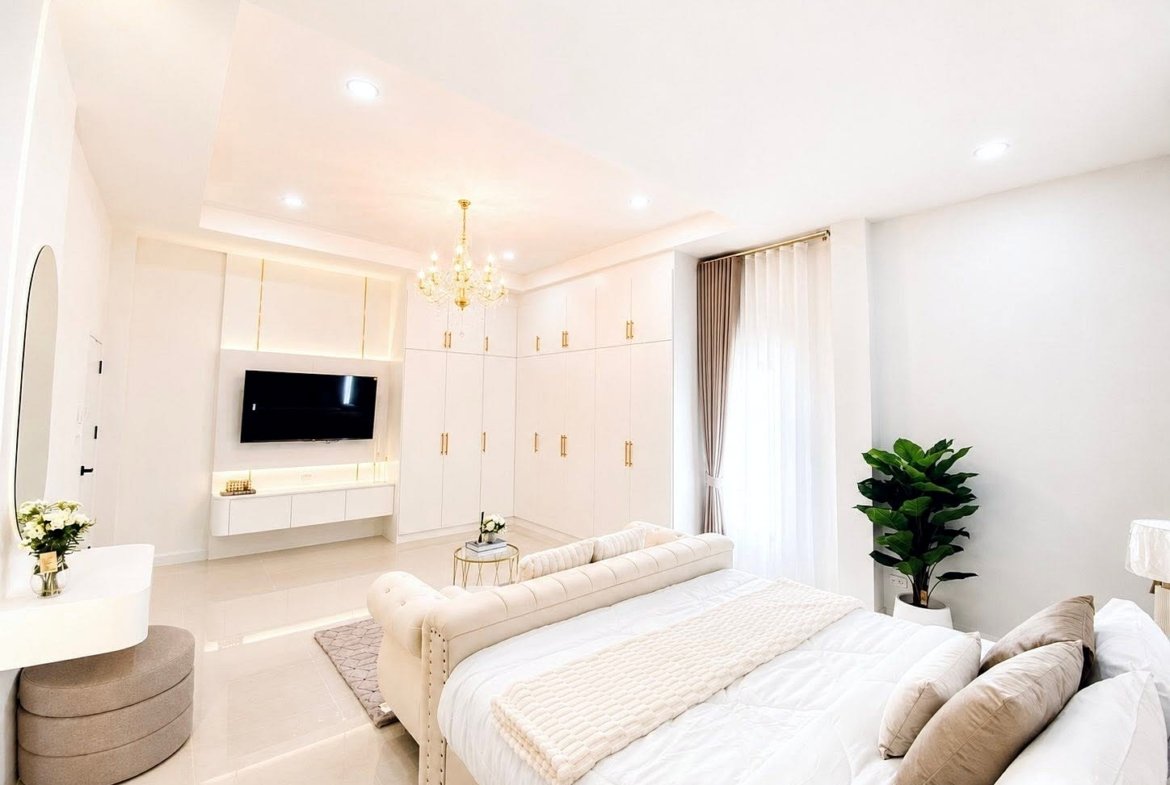 Bright all-white master bedroom with a chandelier, built-in wardrobes, and a wall-mounted TV.