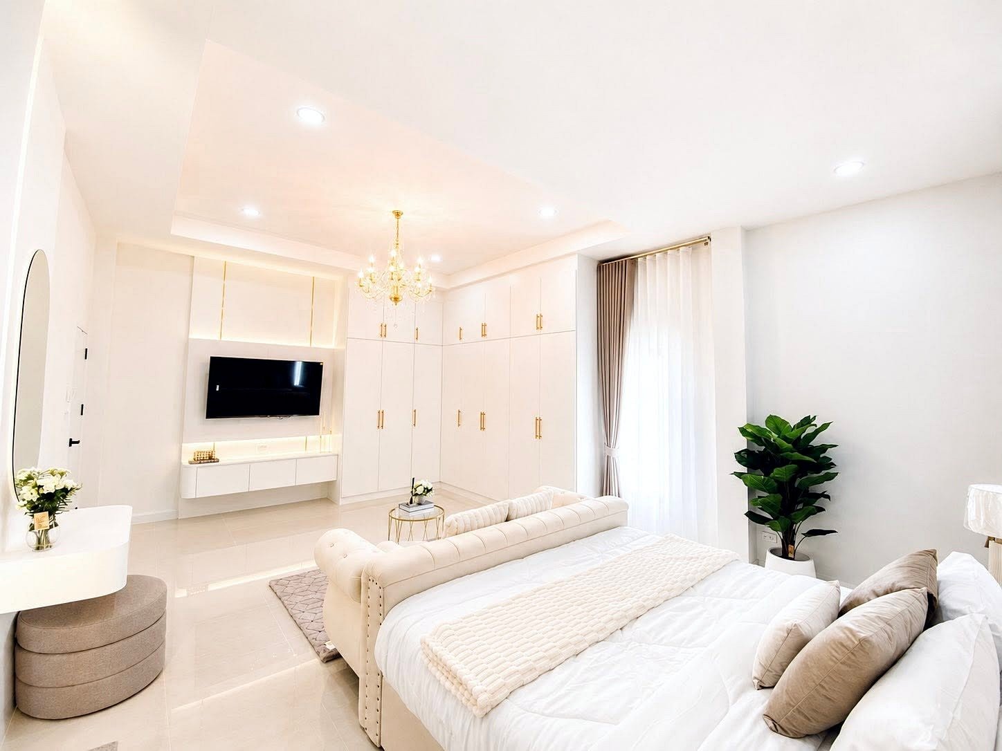 Bright all-white master bedroom with a chandelier, built-in wardrobes, and a wall-mounted TV.
