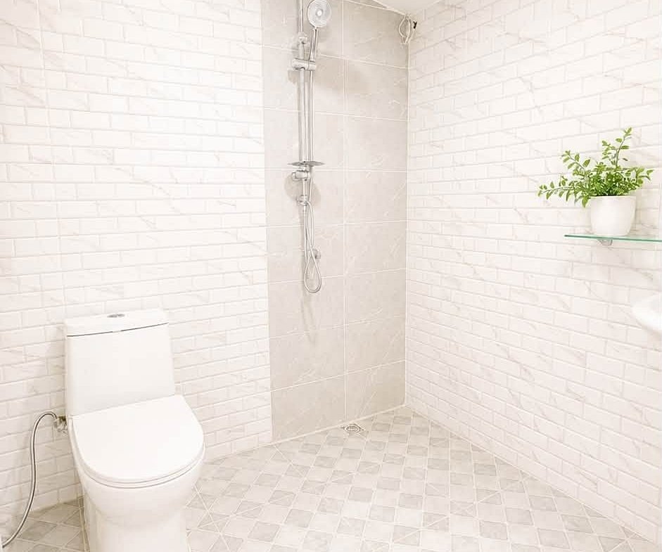Bright white bathroom with a toilet and a walk-in shower, white brick walls, tiled floor, and a potted plant on a glass shelf.