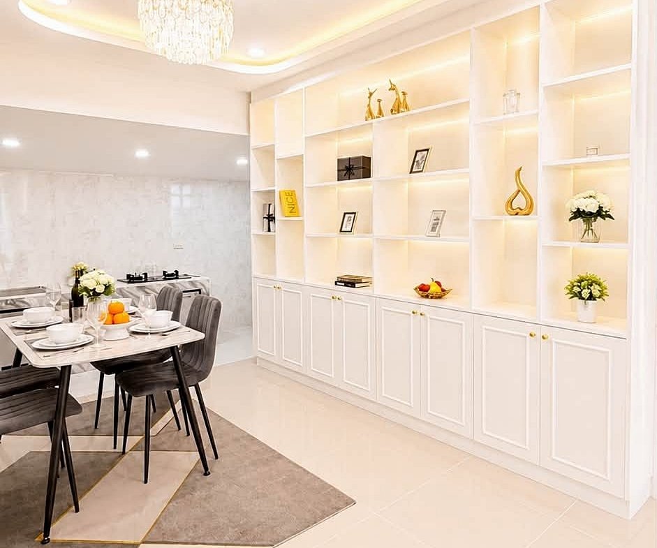 Bright modern dining room with a white built-in shelf wall, crystal chandelier, and a table set for six. Includes a fruit bowl and flowers on the table and soft warm lighting.