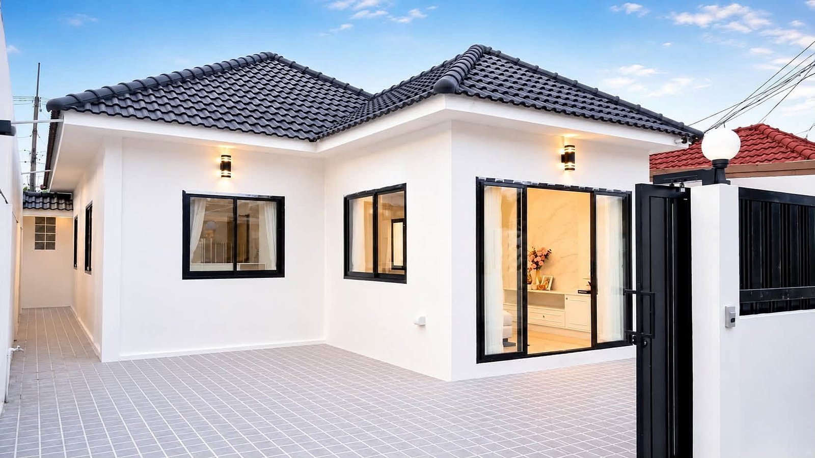 Pattaya_Rungruang_Sale_Dlh579_2 Modern white, single-story home with black-framed windows, gray tiled roof, and an open black gate in a paved courtyard.