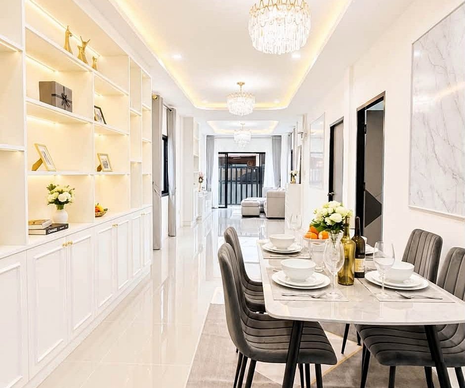 Bright modern dining room with a marble table set for six, white shelves on the left, and crystal chandeliers along a glossy hallway.