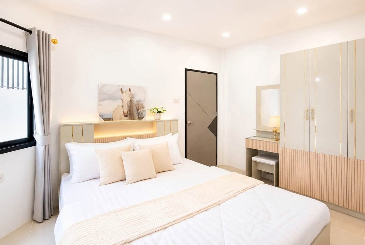 Bright modern bedroom with a large white bed, beige cushions, pale headboard, and a wardrobe along the right side.