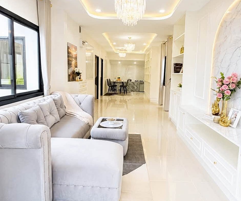 Bright open-concept living room with white sofa, gray rug, and gold chandelier; dining area visible in back.