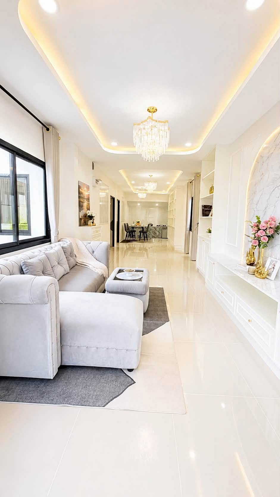 Pattaya_Rungruang_Sale_Dlh579_8 Bright open-concept living room with white sofa, gray rug, and gold chandelier; dining area visible in back.