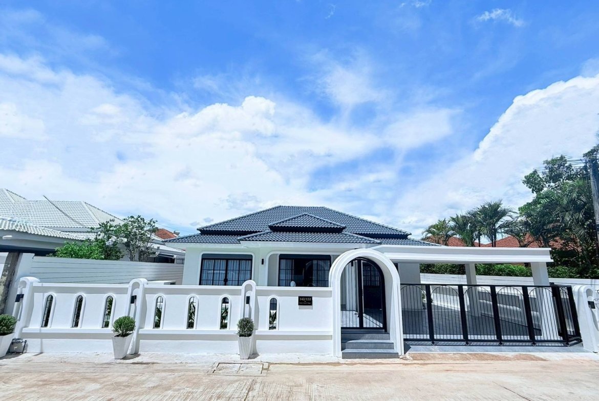 White villa with a decorative arched gate, black fence, and potted shrubs along a white wall under a blue sky.