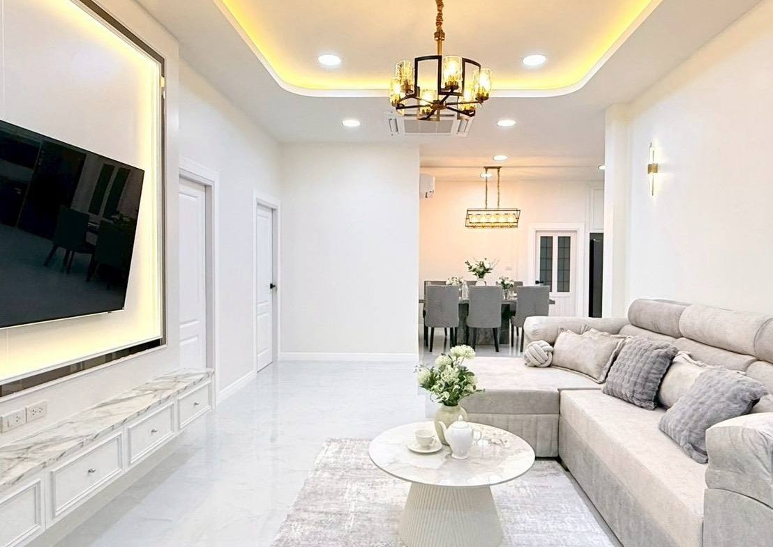 Bright, modern living room with a gray sectional, white marble coffee table, and wall-mounted TV; dining area visible in the back with a chandelier and warm ceiling lights.