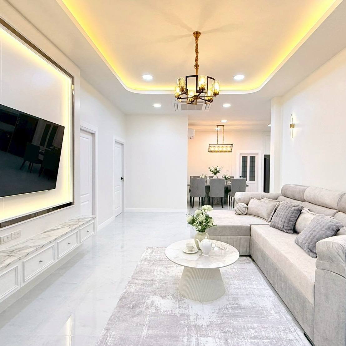 Pattaya_Tropical_2_Village_Dlh535_10 Bright, modern living room with a gray sectional, white marble coffee table, and wall-mounted TV; dining area visible in the back with a chandelier and warm ceiling lights.