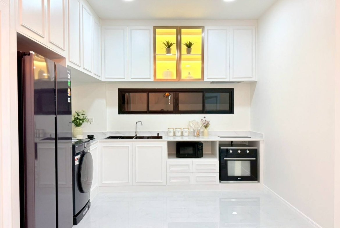 Modern white kitchen with a side-by-side stainless fridge, washing machine, and white cabinets surrounding a marble-like floor.