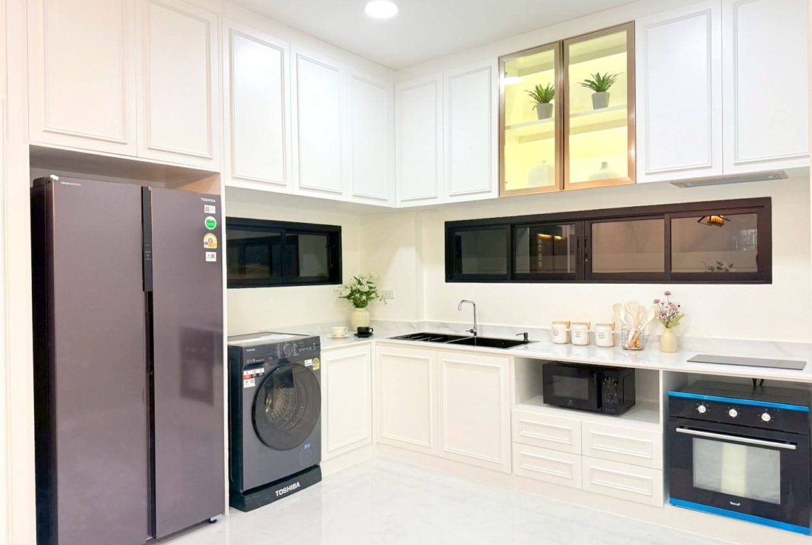 Bright white kitchen with a tall gray side-by-side refrigerator, front-loading washing machine, and white cabinets along the walls near a black-framed windowed counter edge.