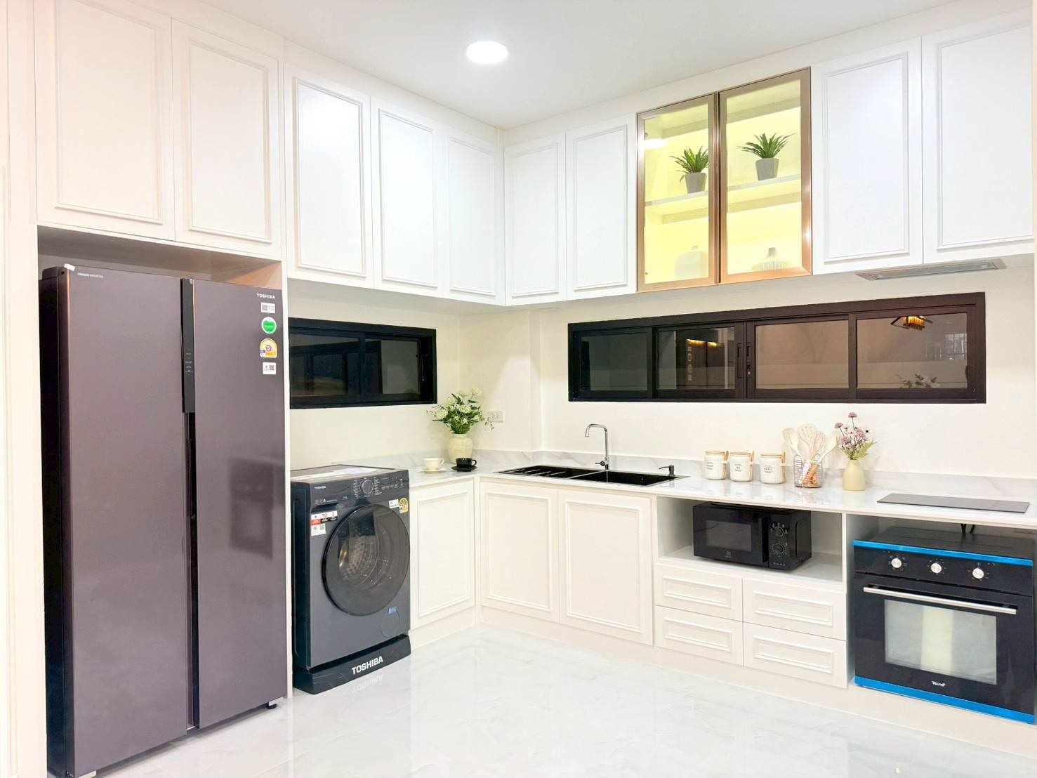 Pattaya_Tropical_2_Village_Dlh535_12 Bright white kitchen with a tall gray side-by-side refrigerator, front-loading washing machine, and white cabinets along the walls near a black-framed windowed counter edge.