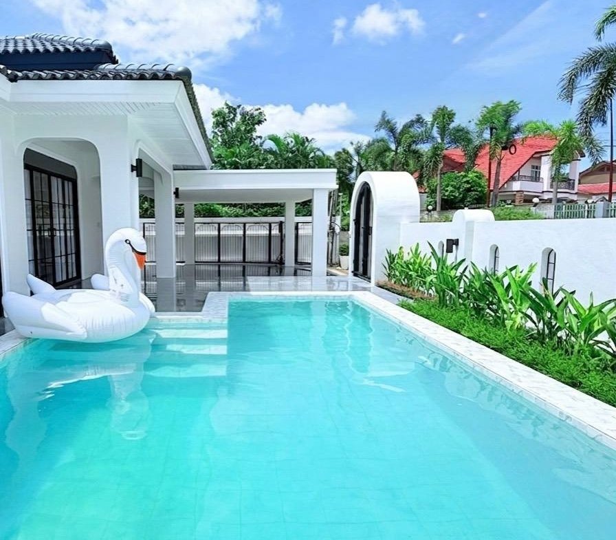 White modern house with a rectangular turquoise pool, two white swan pool inflatables, and a covered patio area; bright blue sky and tropical plants in the background.