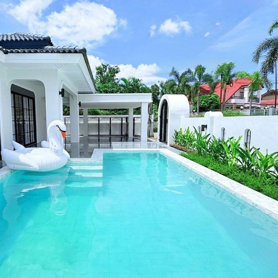 Pattaya_Tropical_2_Village_Dlh535_13 White modern house with a rectangular turquoise pool, two white swan pool inflatables, and a covered patio area; bright blue sky and tropical plants in the background.
