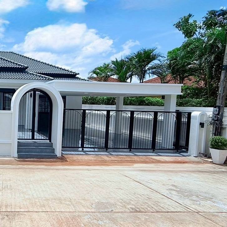 Modern gated entrance to a house: white arched doorway with gray steps, black vertical-bar gate, and a paved driveway; palm trees in the background.