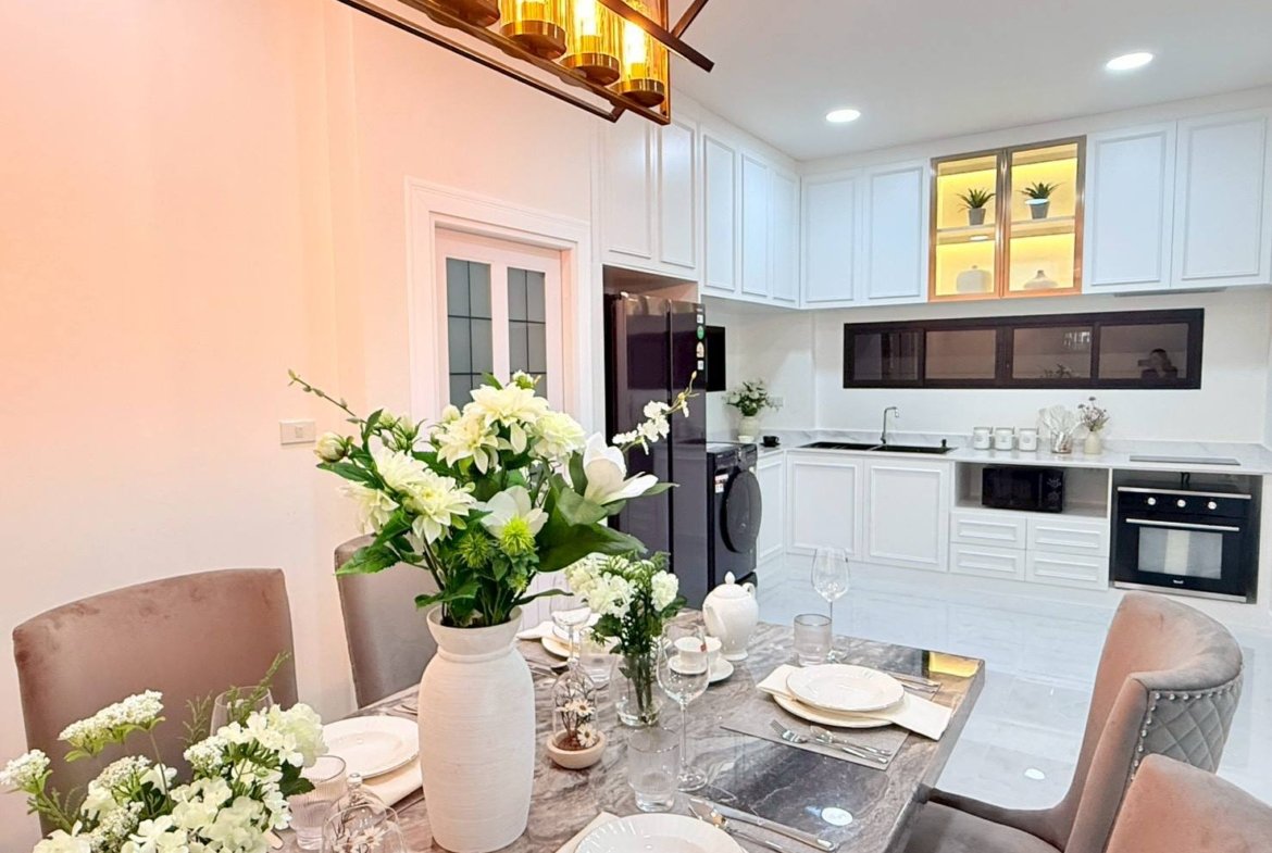 Dining table set for six in a bright, modern kitchen with a tall white vase of white flowers as the centerpiece and a gold-toned chandelier above.