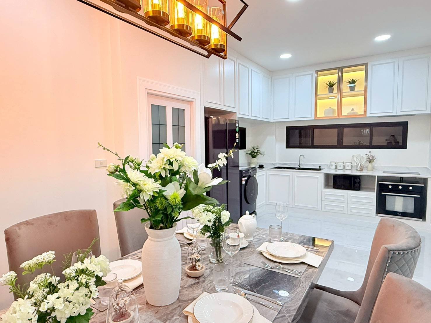 Pattaya_Tropical_2_Village_Dlh535_4 Dining table set for six in a bright, modern kitchen with a tall white vase of white flowers as the centerpiece and a gold-toned chandelier above.