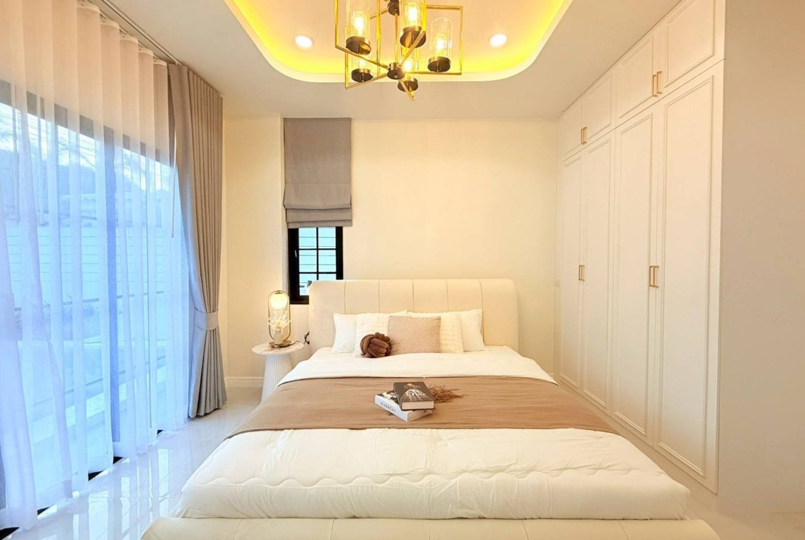 Bright modern bedroom with a white upholstered bed, beige headboard, and built-in white wardrobes on the right; sheer curtains over large windows.